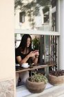 Sorridente donna asiatica chatta con il suo telefono in una caffetteria accanto alla finestra — Foto stock
