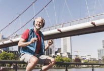 Happy athletic mature man with headphones sitting at the riverside in the city — Stock Photo