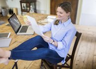 Donna sorridente seduta alla scrivania a casa con i piedi alzati e il documento di lettura — Foto stock