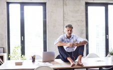 Uomo d'affari maturo con smartphone e cuffie seduto a piedi nudi sulla scrivania in ufficio — Foto stock