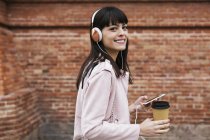 Retrato de mulher sorridente com café, celular e fones de ouvido contra parede de tijolo — Fotografia de Stock