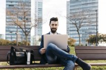 Businessman sitting on bench outside office building and using laptop — Stock Photo