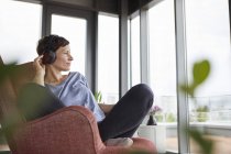Mulher sentada em poltrona em casa ouvindo música com fones de ouvido — Fotografia de Stock