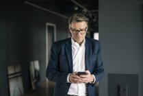 Зрелый бизнесмен, стоящий в офисе с помощью мобильного телефона — стоковое фото