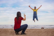 Femme prenant une photo smartphone de l'homme heureux sautant sur la plage — Photo de stock