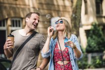 Couple heureux avec café pour aller souffler des bulles de savon dans la ville — Photo de stock