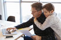 Businessman using laptop at desk in office with son embracing him — Stock Photo