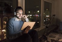 Businessman sitting on window sill in office at night using tablet and headphones — Stock Photo