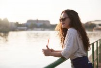 Усміхаючись молода жінка з винос напій насолоджуючись заходом сонця на березі річки — стокове фото