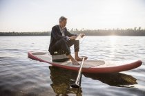 Uomo d'affari seduto sul paddleboard sul lago e utilizzando tablet con auricolari — Foto stock
