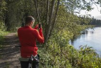 Горный велосипедист, стоящий у озера и фотографирующий со смартфоном — стоковое фото