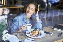 Усміхнена молода жінка насолоджується млинцями та кавою в кафе — стокове фото