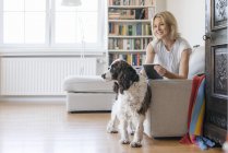 Усміхнена зріла жінка з собакою лежачи на дивані вдома і тримає планшет — стокове фото