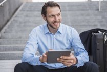 Smiling businessman with rolling suitcase sitting on stairs using tablet — Stock Photo