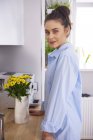 Jeune femme à la maison, debout dans la cuisine à table avec des fleurs jaunes et posant sur la caméra — Photo de stock