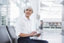 Senior businesswoman sitting in waiting area with tablet and looking around — Stock Photo