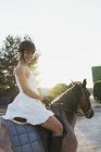 Portrait of smiling woman in dress and helmet sitting on horse in backlit — Stock Photo