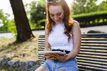 Смеющаяся рыжеволосая женщина, сидящая на скамейке в парке и использующая цифровой планшет — стоковое фото