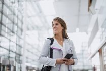Retrato de jovem empresária sorridente com telefone celular no aeroporto olhando ao redor — Fotografia de Stock