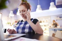 Sourire jeune femme travaillant dans un café à l'aide d'un ordinateur portable et d'un téléphone portable — Photo de stock