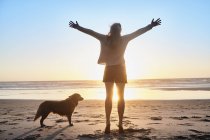 Португалія, Алгарве, жінка з собакою, що піднімають руки на пляжі на заході сонця — стокове фото