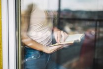 Primo piano di una giovane donna che legge un libro dietro il vetro della finestra — Foto stock