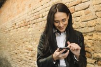 Усміхнена молода жінка використовує мобільний телефон на цегляній стіні — стокове фото