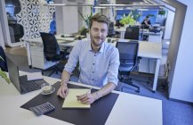Portrait of smiling businessman sitting at desk in open-plan office — Stock Photo
