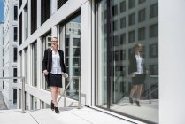 Femme d'affaires souriante avec journal marchant devant un immeuble de bureaux — Photo de stock