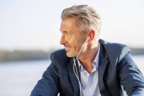Homme d'affaires souriant écoutant de la musique avec des écouteurs au lac — Photo de stock