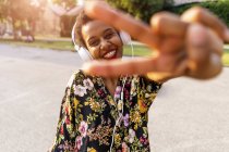 Happy fashionable young woman with headphones outdoors at sunset making victory gesture — Stock Photo