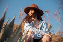 Улыбающаяся молодая женщина в шляпе сидит на агаве в сельской местности — стоковое фото