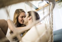Feliz pareja cariñosa besándose en silla colgante en casa - foto de stock