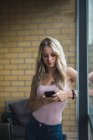 Giovane donna bionda che controlla il cellulare alla finestra — Foto stock
