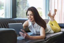 Smiling woman lying on couch at home using a tablet — Stock Photo