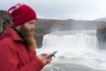 Island, Norden von Island, junger Mann mit Smartphone, Wasserfall im Hintergrund — Stockfoto