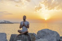 España. Hombre meditando durante el amanecer en la playa rocosa - foto de stock