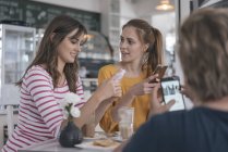 Two girlfriends meeting in a coffee shop, using smartphones — Stock Photo