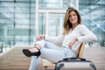 Sorridente giovane donna d'affari seduta all'aperto con valigia guardarsi intorno — Foto stock