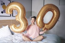 Mujer feliz sosteniendo globos de oro en la mañana de cumpleaños en la cama - foto de stock