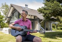 Smiling mature man sitting in garden of his home playing guitar — Stock Photo