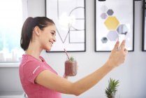 Sorridente giovane donna con frullato scattare selfie con smartphone in cucina — Foto stock