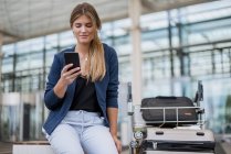 Lächelnde junge Geschäftsfrau sitzt draußen mit Handy und Koffer — Stockfoto