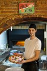 Ritratto di pizzaiolo sorridente che tiene una pizza in cucina — Foto stock