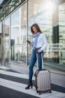 Улыбающаяся молодая деловая женщина с чемоданом, смотрящая на смартфон — стоковое фото