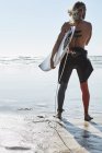 Homme sans chemise confiant debout sur la plage avec planche de surf — Photo de stock