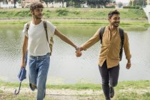 Ritratto di giovane coppia gay con zaini che camminano mano nella mano sul lungofiume — Foto stock