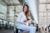 Giovane donna d'affari sorridente seduta sui bagagli con il cellulare che si guarda intorno — Foto stock