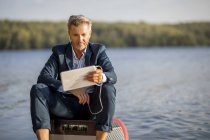 Retrato de hombre de negocios relajado con auriculares y tableta sentado en el paddleboard en el lago - foto de stock