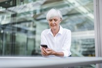 Empresária sênior apoiada em trilhos na cidade e usando telefone celular — Fotografia de Stock